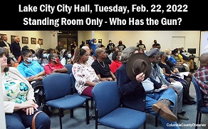 Photo: Lake City City Hall with standing room only with one attendee covering his face with his hat. Caption: Lake City, City Hall, February 22, 2022. Standing room only - who has the gun?